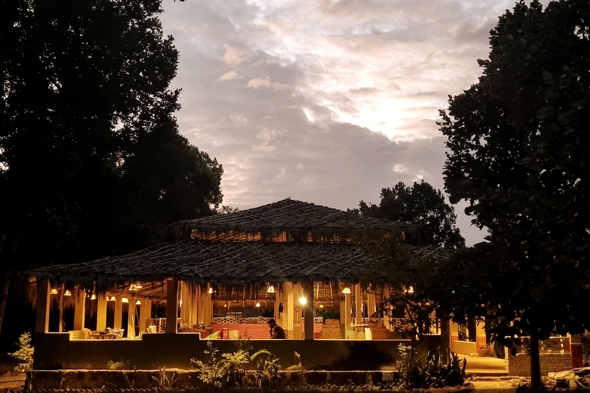 An open-air dining area is showcased under a thatched roof, surrounded by lush greenery. Soft lighting creates a warm ambiance, while the night sky and clouds form a serene backdrop. A cozy seating arrangement invites guests to enjoy meals amid nature.