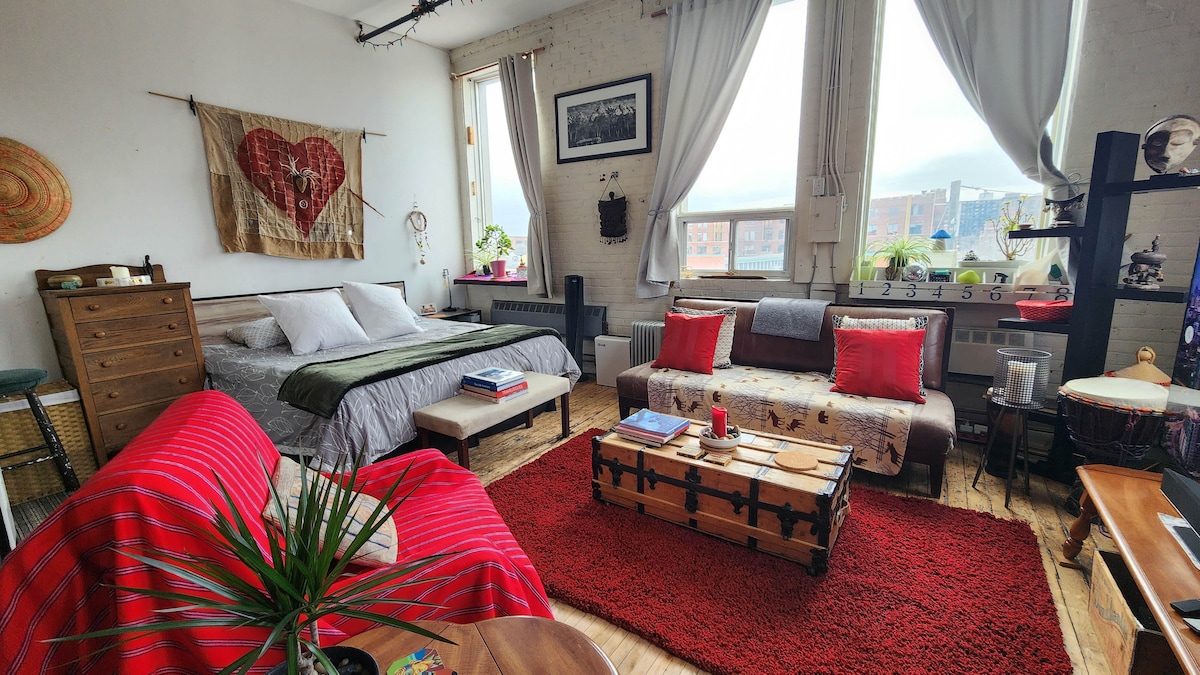 A spacious open-concept loft features large windows that invite natural light. A cozy seating area is arranged with comfortable couches on a vibrant red rug. A wooden coffee table with storage sits in the center, while a queen bed is adorned with neutral bedding and decorative pillows.