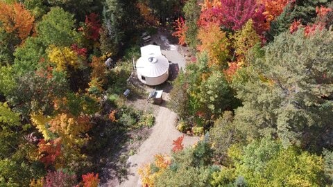 An aerial view captures a white yurt nestled among vibrant autumn foliage. A winding path leads to the yurt, surrounded by trees displaying a spectrum of red, orange, and yellow leaves. Natural landscaping complements the secluded setting.