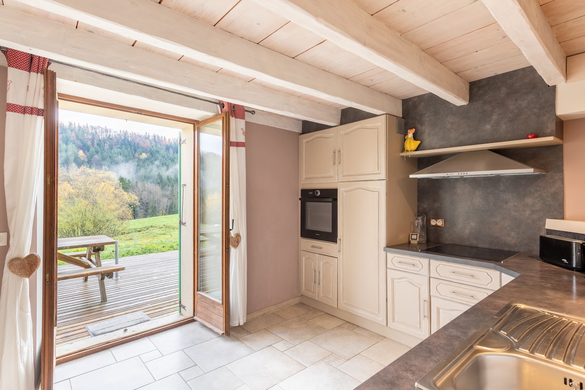A bright kitchen space features wooden beams and light-colored cabinetry. An oven and stove are visible, along with a stainless steel sink and countertop. Double doors open to a serene outdoor view of greenery, inviting a connection with nature.