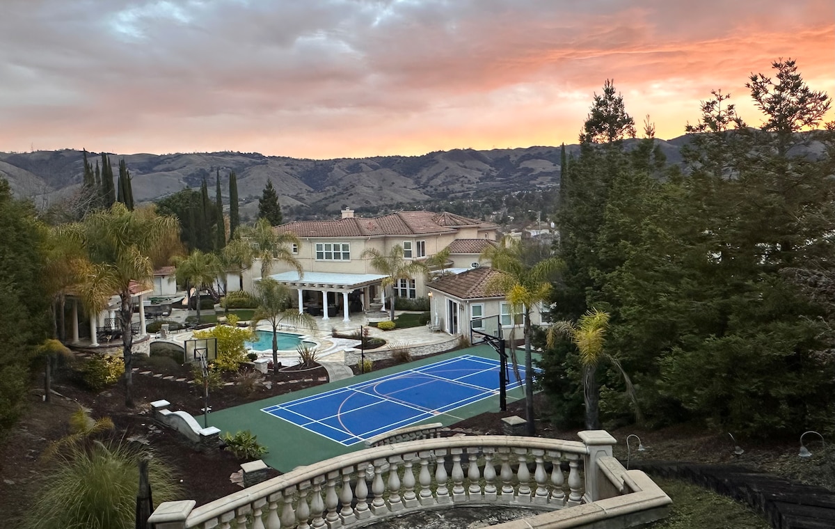 An expansive view showcases the outdoor amenities, including a well-maintained basketball court and lush landscaping. The backdrop features rolling hills under a colorful sunset sky, with a spacious residence and patio area visible in the distance surrounded by palm trees.