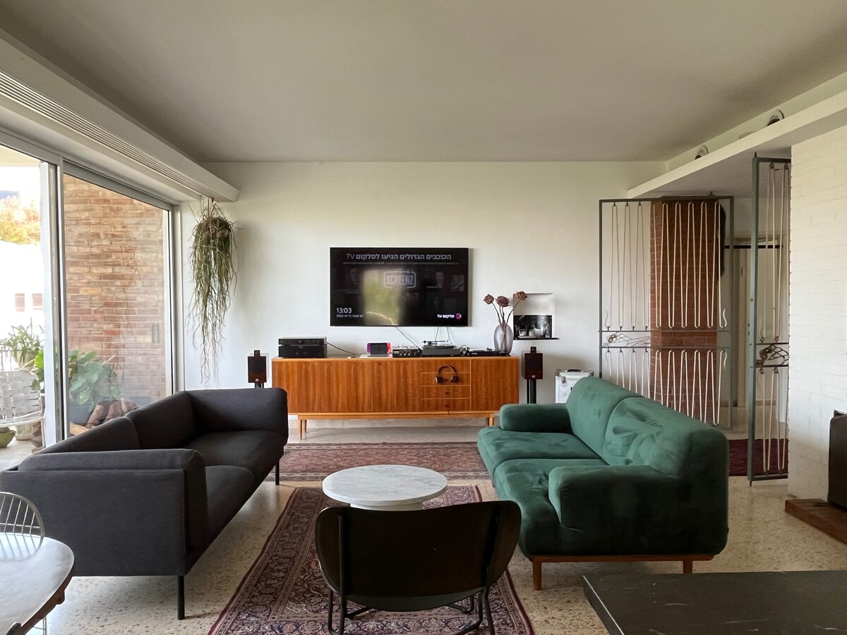 A modern living room is displayed with a green sofa and a dark couch arranged around a round coffee table. A wooden media console sits against the wall, with a television mounted above it. Large glass doors invite natural light, while a rug adds warmth and texture.