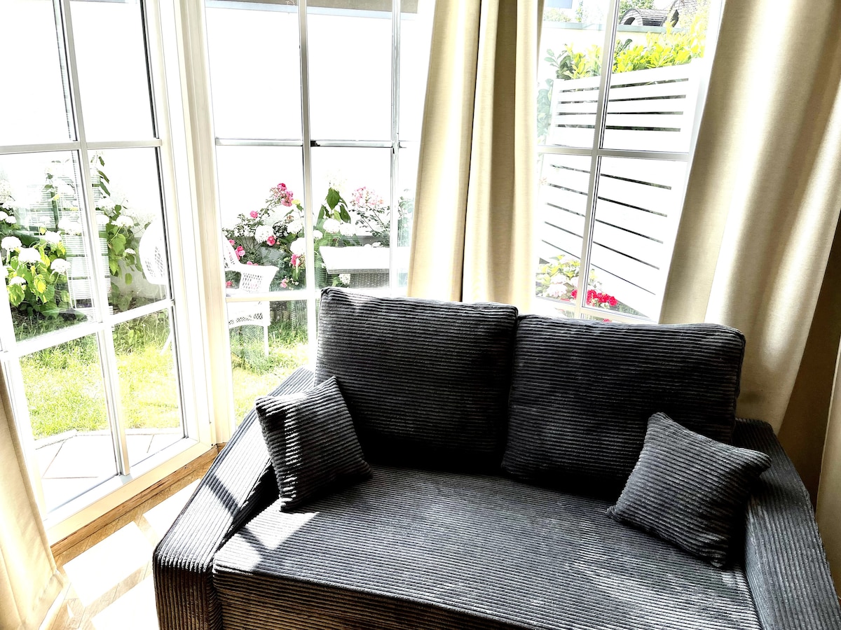 A comfortable, gray-striped sofa is positioned near large windows that allow natural light to fill the space. A view of flowering plants and green grass can be seen through the windows, creating a serene setting.
