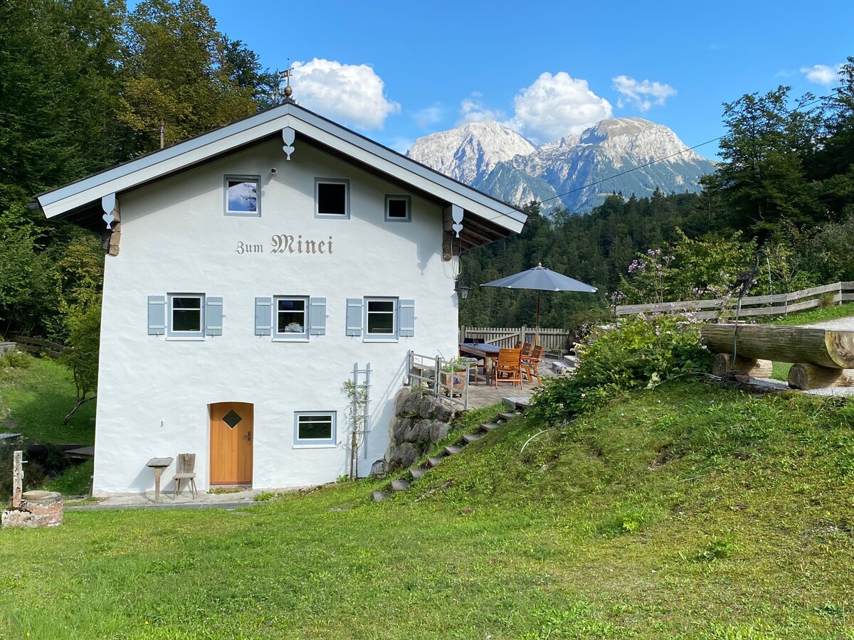 A charming two-story house with a white façade is nestled in a green landscape. A wooden door is visible, along with a patio area featuring outdoor seating and an umbrella. Majestic mountains rise in the background under a clear blue sky.