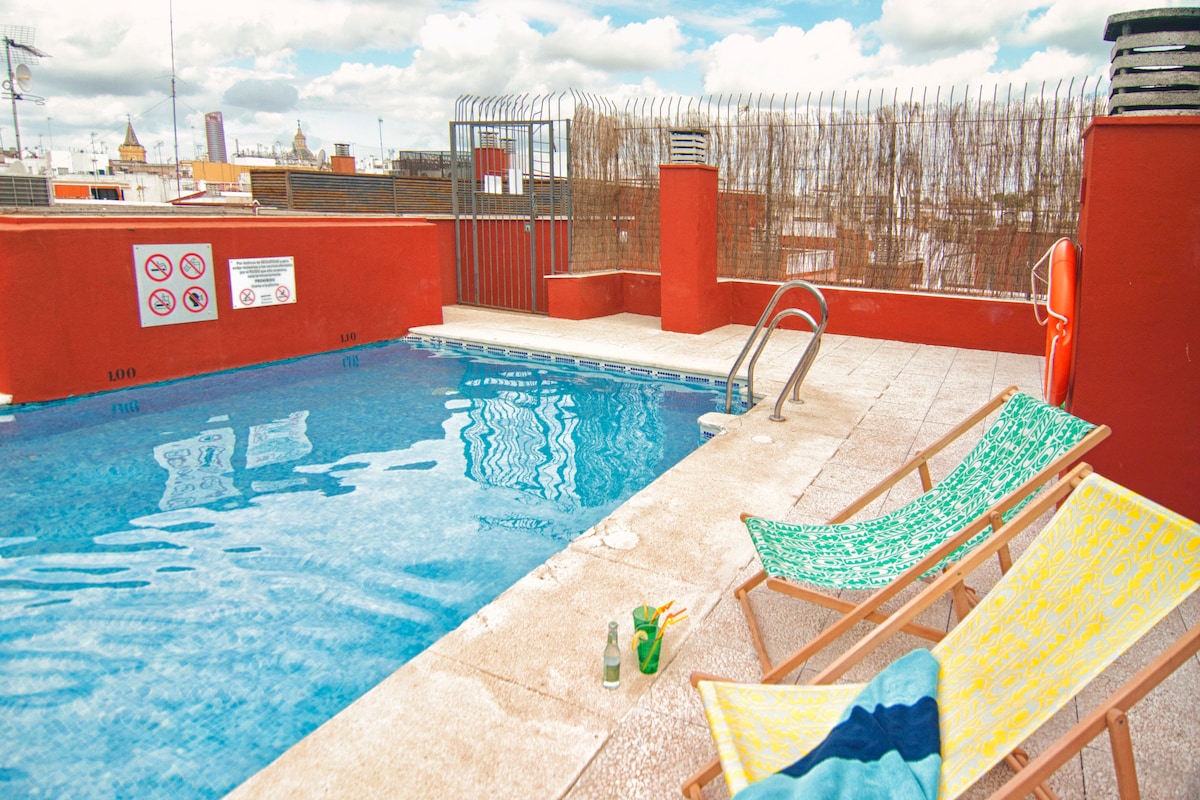 A community rooftop swimming pool is surrounded by a low barrier and features two lounge chairs positioned nearby. The pool’s clear water reflects the sky, while a vibrant orange wall adds a touch of warmth to the space. Access to the pool is available during the summer season.