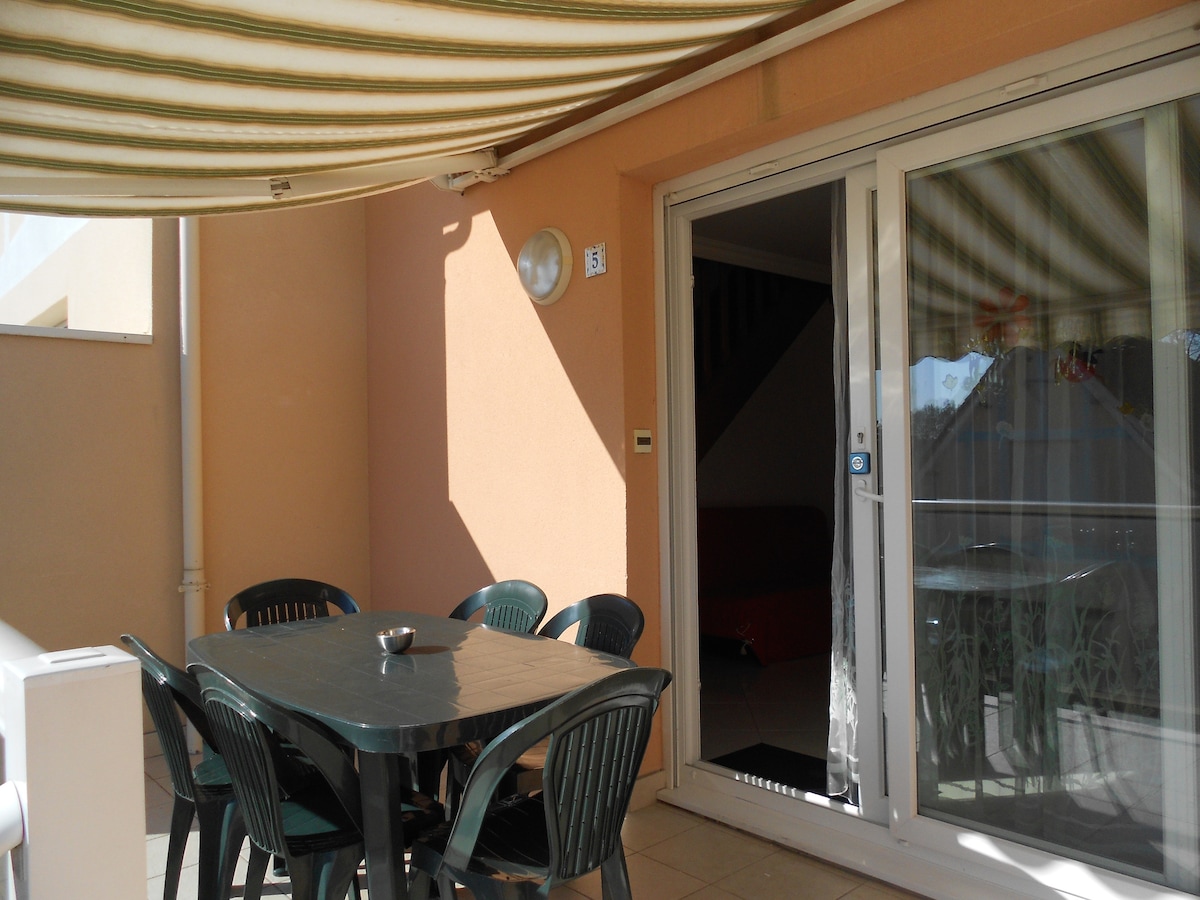 A covered terrace features a dark table surrounded by several green chairs. Natural light brightens the space, with a door leading indoors visible in the background. The walls are finished in a warm tone, complementing the outdoor setting.