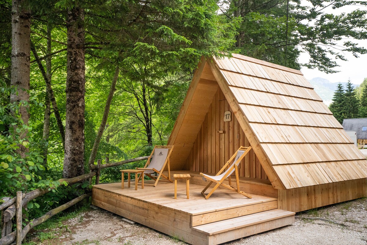 A wooden villa stands amidst lush greenery, featuring a sloped roof and a welcoming wooden deck. Two deck chairs are positioned on the deck, inviting relaxation in the natural surroundings. The area is surrounded by tall trees, enhancing the tranquil atmosphere.