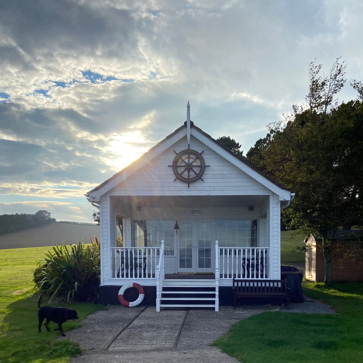 A charming white cottage is presented with a nautical theme, featuring a ship's wheel above the entrance. The porch is framed by a railing and plants, while sunlight casts a warm glow. A dog is seen on the grassy path beside the house.