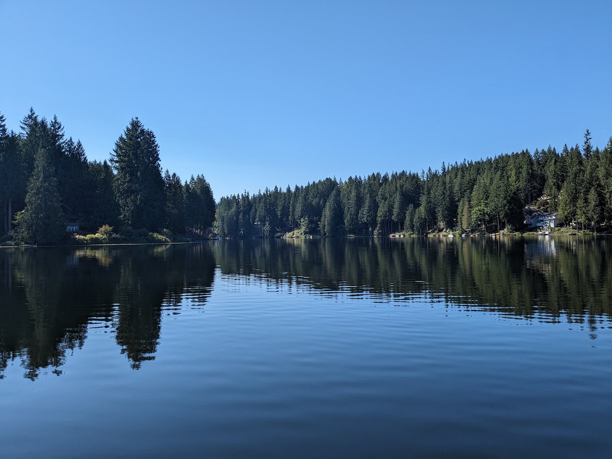 A serene view of Ames Lake reflects the clear blue sky and surrounding evergreen trees. The calm water creates a mirror-like surface, enhancing the peaceful atmosphere. The shoreline reveals a mix of natural vegetation and structures, inviting exploration and relaxation.