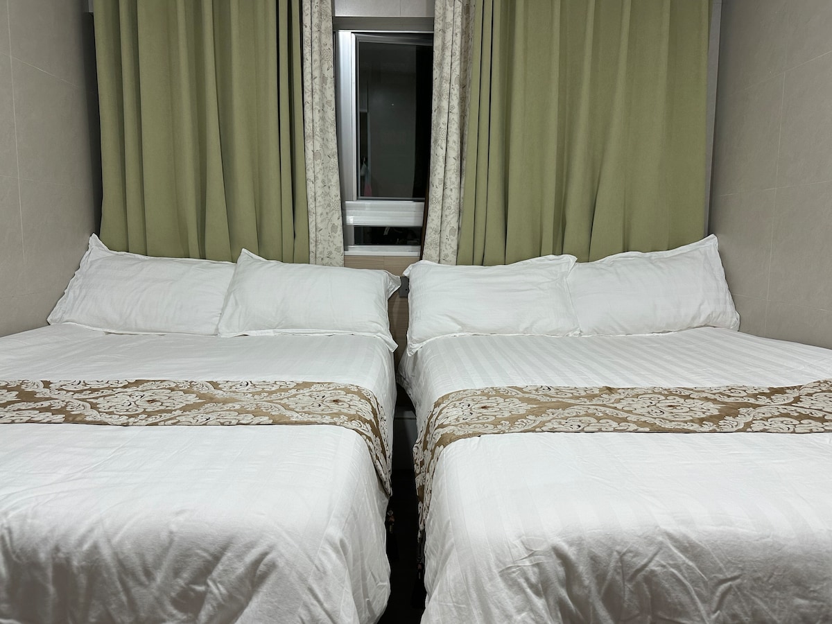 Two spacious beds are positioned close together, adorned with white linens and brown decorative throws. Light green curtains frame a nearby window, allowing natural light to filter in softly.