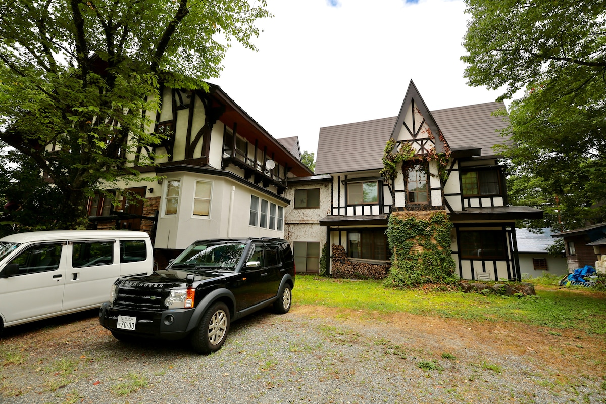 A charming two-story building showcases a blend of architectural styles with a sloped roof and decorative elements. Surrounded by greenery, it features multiple windows and a driveway with parked vehicles. The natural setting provides a serene environment.