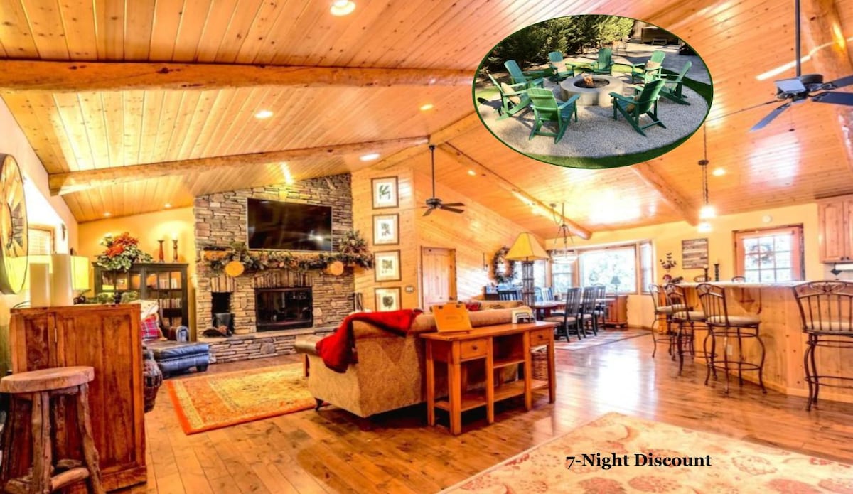 A spacious living area features vaulted ceilings with exposed wooden beams. A large stone fireplace emphasizes a cozy atmosphere, while a comfortable seating arrangement invites relaxation. A smart TV is mounted above the fireplace, and the dining area is visible in the background.