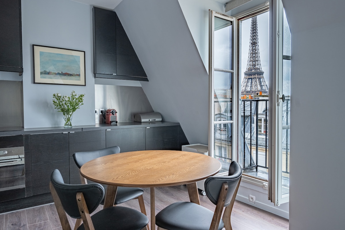 A modern kitchen area features a round wooden table surrounded by four black chairs. Natural light enters through an open window, revealing a view of the Eiffel Tower. Sleek black cabinetry complements stainless steel appliances.