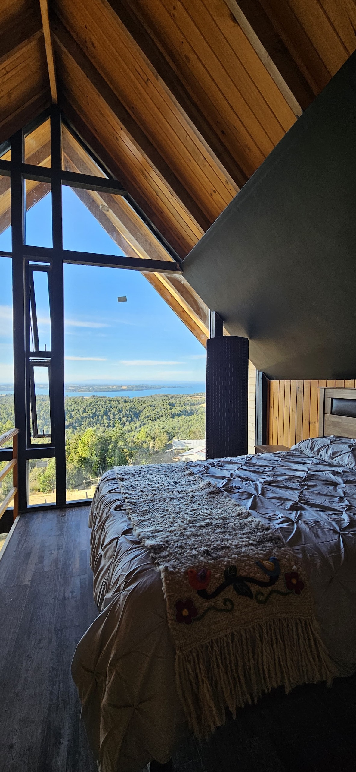 A spacious bedroom features a large window showcasing a panoramic view of the surrounding landscape. The wooden ceiling adds warmth, while the plush bed is adorned with a textured blanket. Natural light fills the room, creating an inviting atmosphere.