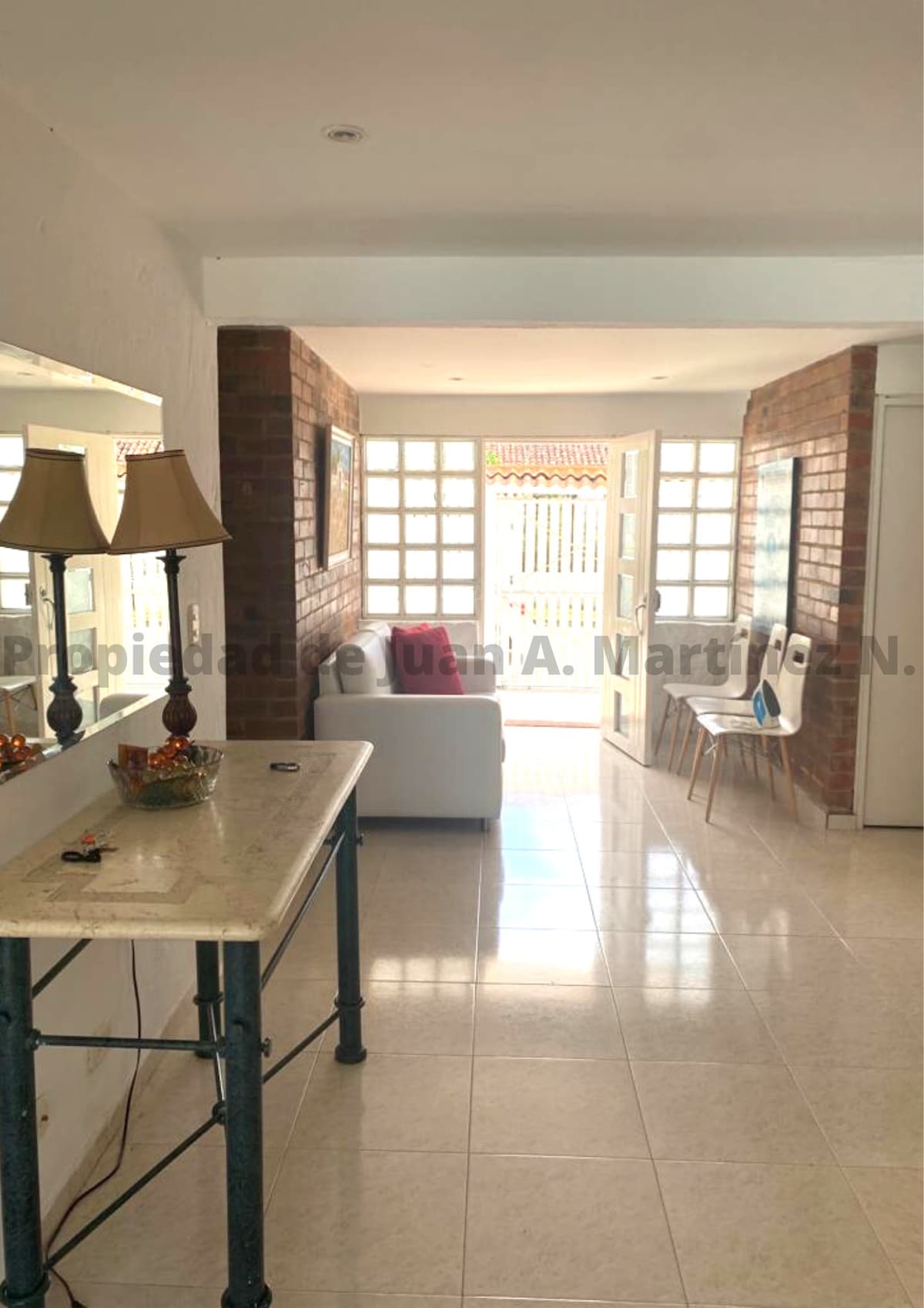 A bright hallway features light-colored tiles and ample natural light streaming through large windows. A white sofa is positioned to one side, while a decorative console table with an ornamental centerpiece stands nearby. The entrance opens to an outdoor area lined with brick accents.