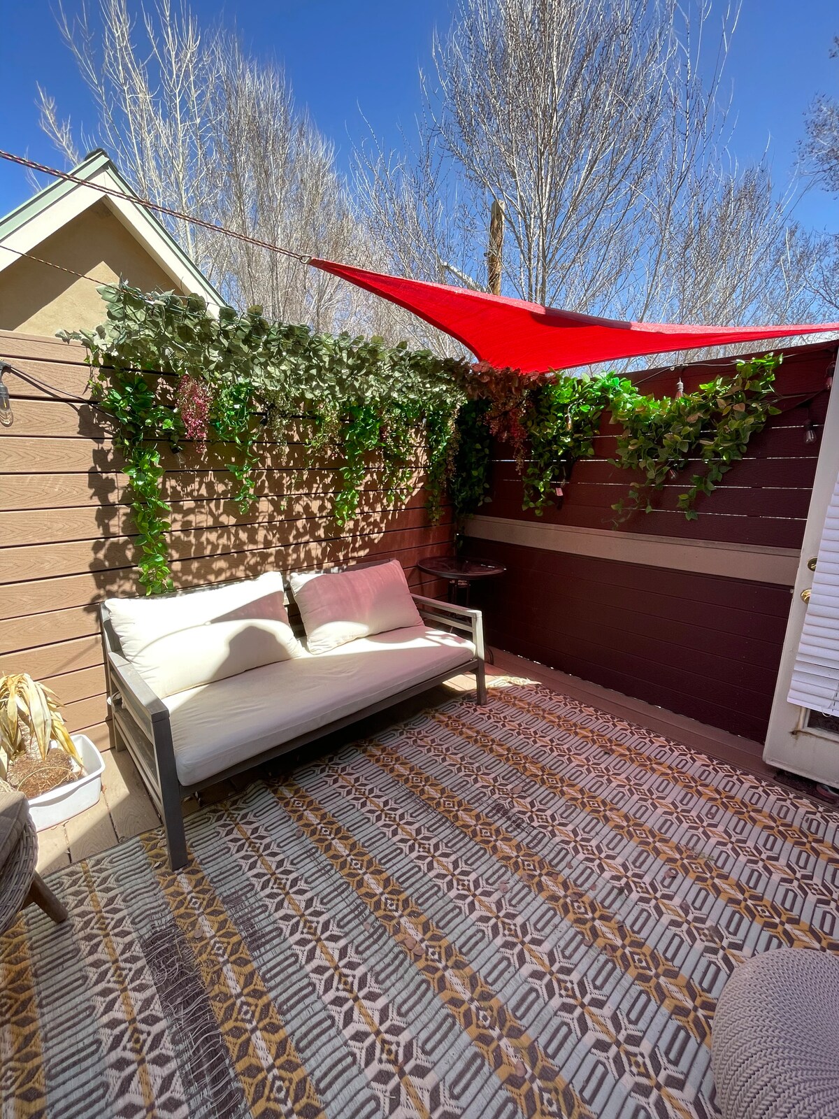 A cozy outdoor patio area is highlighted with a sunshade and surrounded by lush greenery. A light-colored couch offers a comfortable seating option, positioned over a patterned rug that adds texture. Bright blue skies and leafless trees are visible in the background.