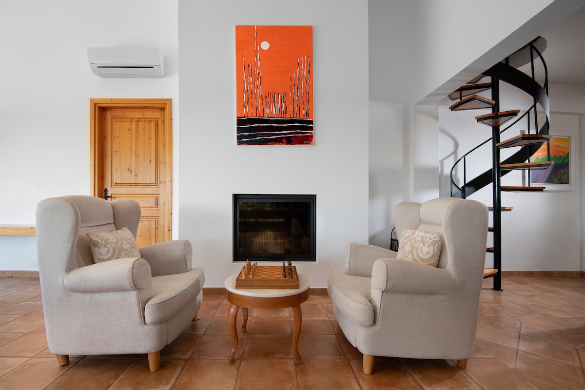 A light and airy living area is presented, featuring two upholstered armchairs positioned beside a wooden coffee table. A modern painting in vibrant orange decorates the wall above a sleek fireplace. A spiral staircase leads to the upper level, enhancing the open feel of the space.
