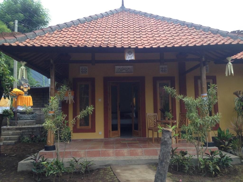 A charming traditional Balinese homestay is presented, featuring a distinctive peaked roof and vibrant yellow exterior. The entrance is flanked by potted plants, and wooden seating is visible on the porch. Steps lead up to the inviting entrance, surrounded by lush greenery.