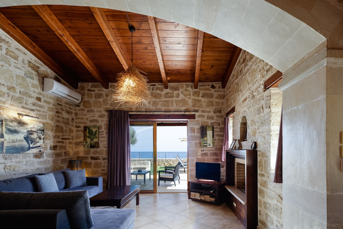 The living area features stone walls and wooden beams, creating a rustic ambiance. A comfortable gray sofa is arranged around a wooden coffee table, while large glass doors open to a private terrace overlooking the sea. Natural light illuminates the space, highlighting its inviting atmosphere.