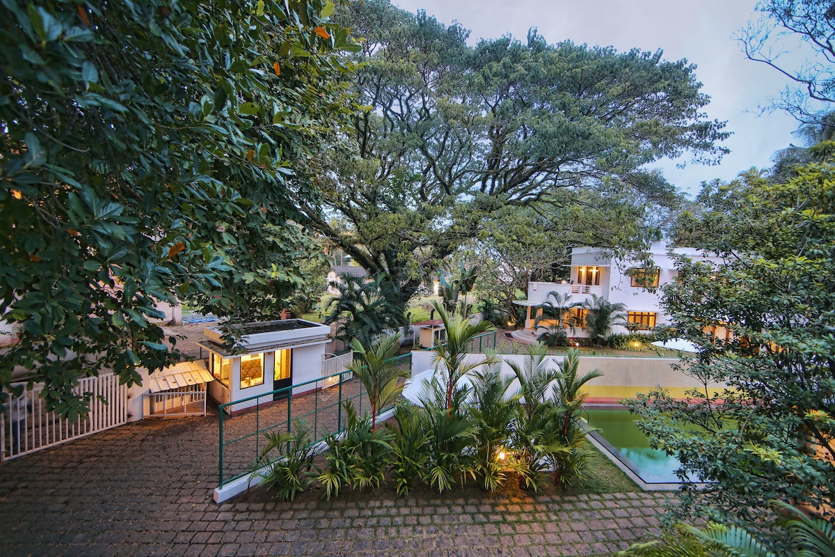 A view of the tranquil garden area features lush greenery, including palm and rain trees. The private swimming pool is visible, framed by tropical plants. The bungalow is partially seen in the background, surrounded by soft evening light.