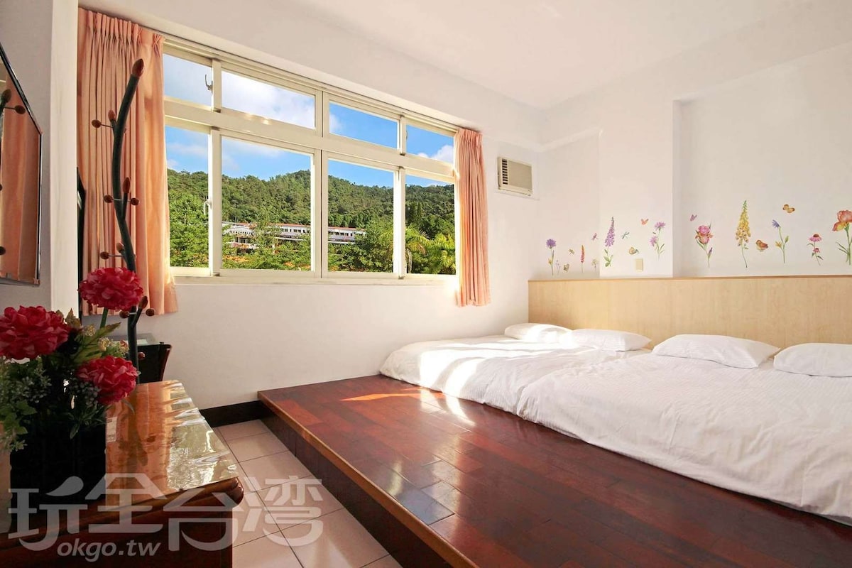 A bright and airy room features large windows showcasing a view of the surrounding greenery. Two spacious beds are positioned on a wooden floor, complemented by floral wall decorations. Soft natural light enhances the inviting atmosphere of the room.