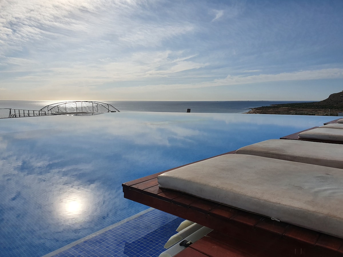 An infinity pool with a wooden deck overlooks the expansive coastline and sea, reflecting the sky above. Comfortable sun loungers are positioned nearby, inviting relaxation while enjoying the serene views.