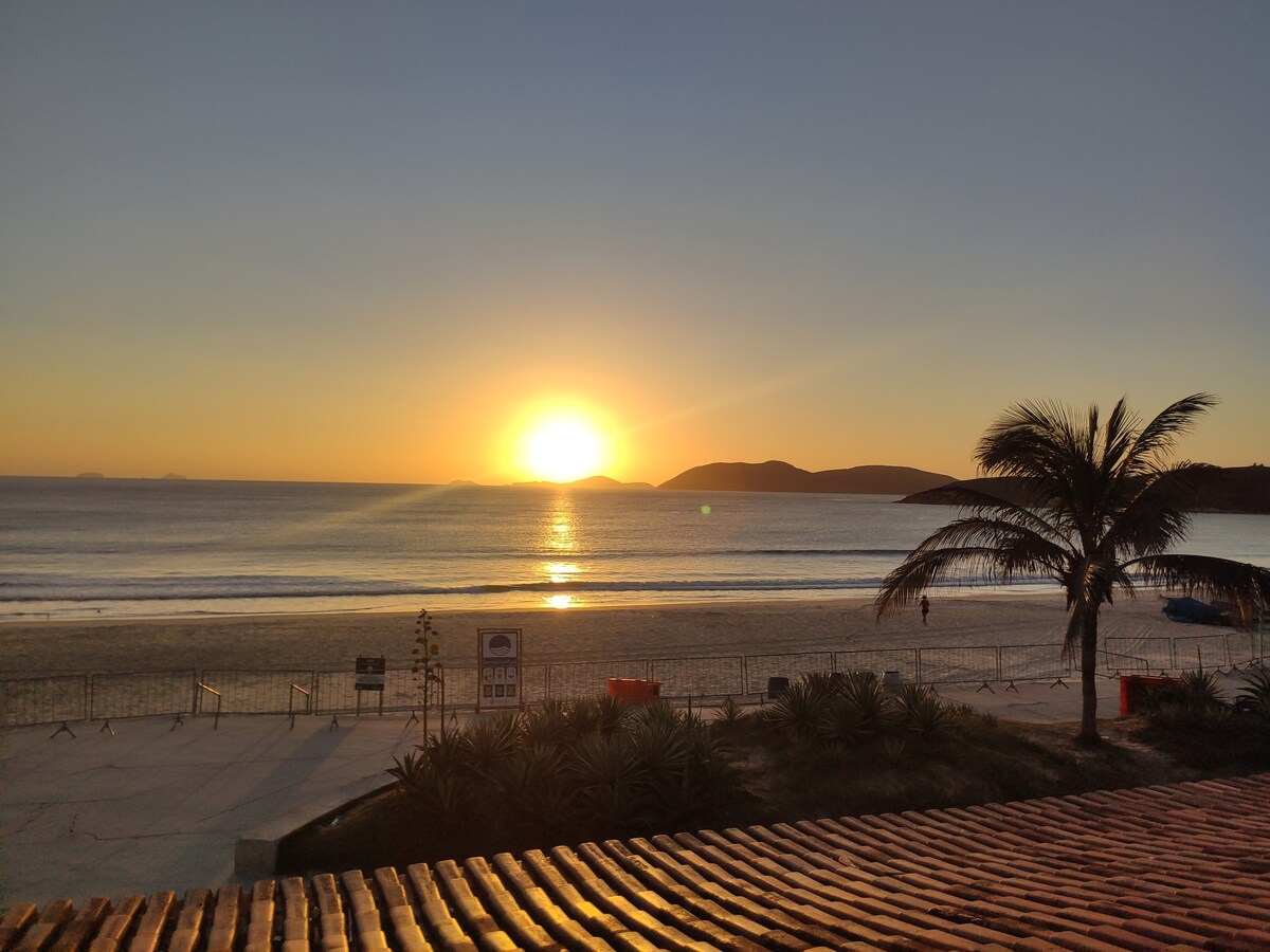 A stunning view captures the sun rising over the tranquil beach, casting golden hues across the water. Silhouetted palm trees frame the scene, while the shoreline is bordered by gentle waves. Coastal hills are visible in the distance, enhancing the serene atmosphere.