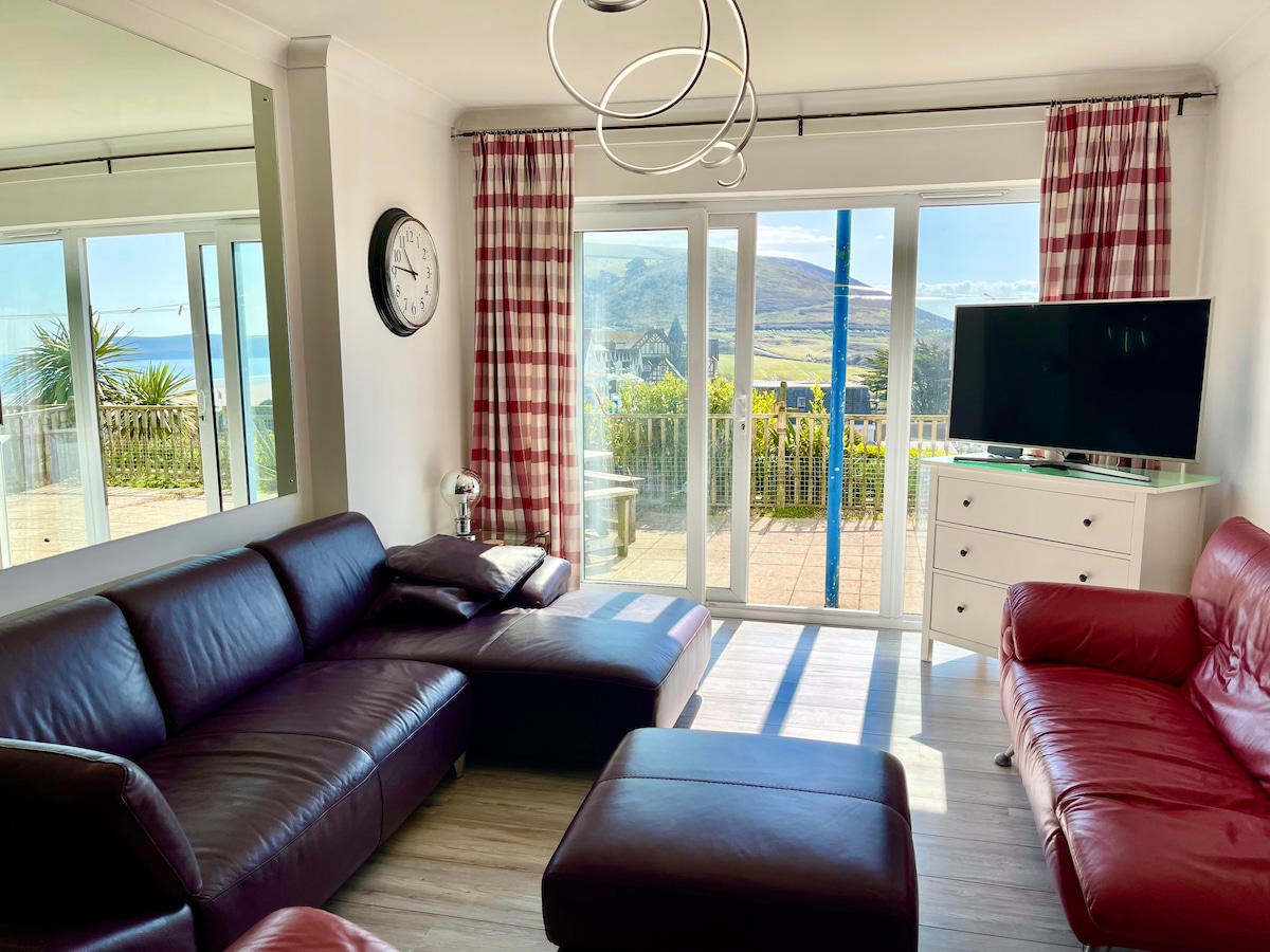 A comfortable living area features a mix of red leather sofas and a large chaise lounge. Natural light enters through expansive glass doors, highlighting a view of the patio and distant hills. Modern furnishings, including a TV and a decorative wall clock, complete the space.