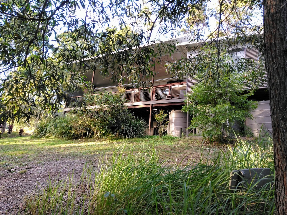 The cottage is surrounded by lush greenery, with large windows allowing ample natural light. A spacious deck extends across the front, providing a shaded outdoor area. Various plants and trees add to the serene setting, showcasing the connection with nature.