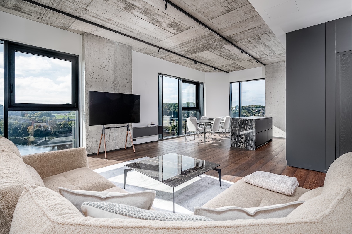 A modern living area features a spacious, light-colored sectional sofa facing a glass coffee table. Large windows offer views of the riverside and natural surroundings, while a sleek dining area is visible in the background, complemented by an elegant kitchen island.