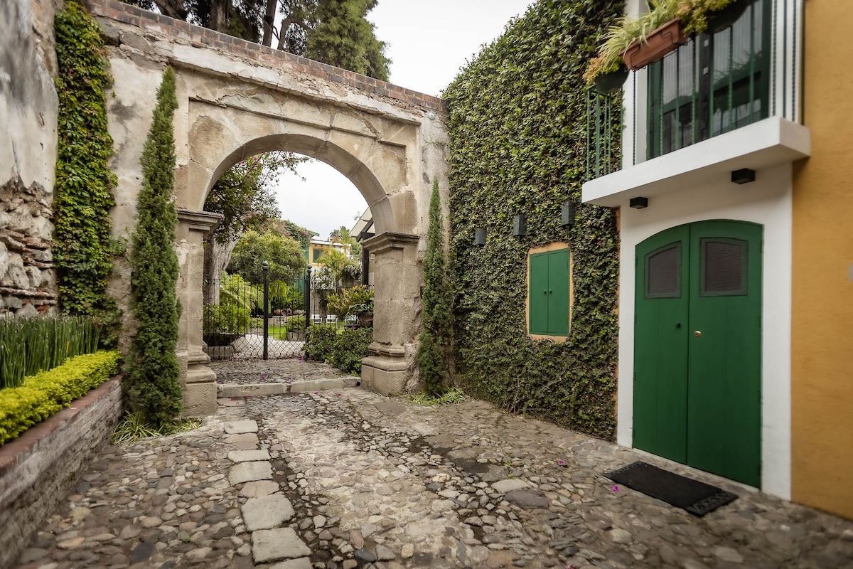 An arched stone entranceway leads into a lush courtyard, where green vines climb the stone walls. Pebbled pathways are bordered by neatly trimmed greenery, creating a serene atmosphere. A vibrant yellow building with two green doors is visible, adding character to the surrounding space.