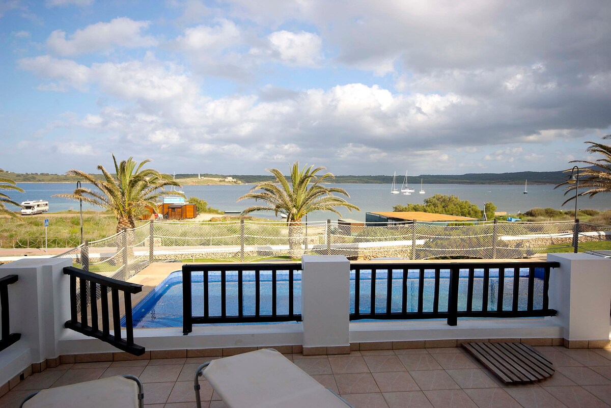 A private terrace overlooks a serene bay, featuring a swimming pool and lined with sun loungers. Palm trees frame the view of the water, where sailboats are anchored. A soft cloud cover enhances the tranquil atmosphere of the surrounding landscape.