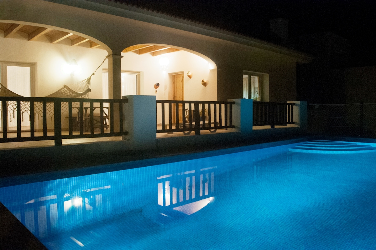An illuminated private pool reflects soft blue tones, enhancing the serene atmosphere. The outdoor area features a covered terrace with several seating options, while warm light spills from the interior, creating a welcoming ambiance. The structure's modern architecture is complemented by a wooden railing.