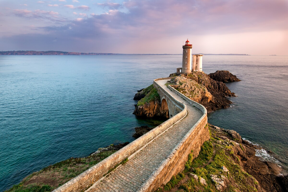 A scenic coastal view features a lighthouse perched on a rocky outcrop, with a winding stone pathway leading towards it. The tranquil sea reflects soft hues from a pastel sky at sunset, enhancing the serene atmosphere of the landscape.