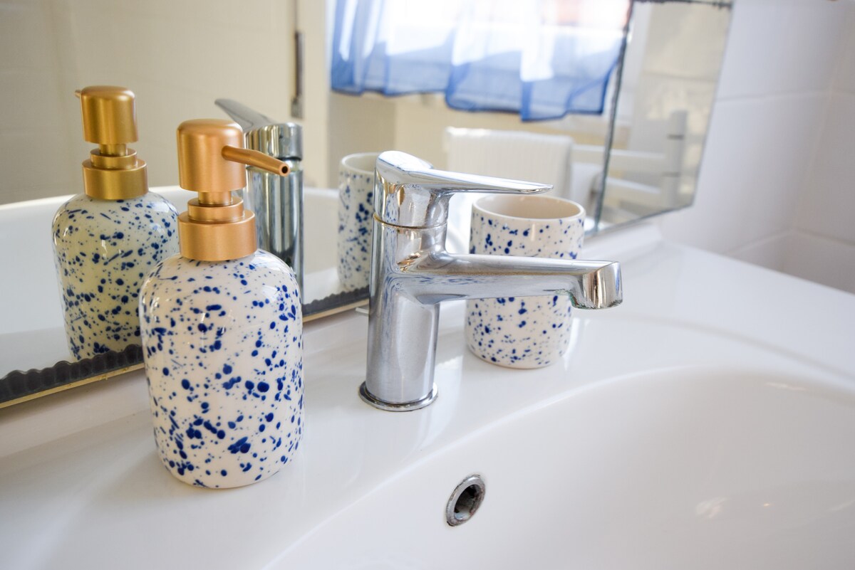 A modern bathroom sink area is presented, featuring a polished chrome faucet and artistic soap dispenser with a speckled blue design. A ceramic cup complements the dispenser. Natural light filters through a sheer curtain, enhancing the clean and functional design.