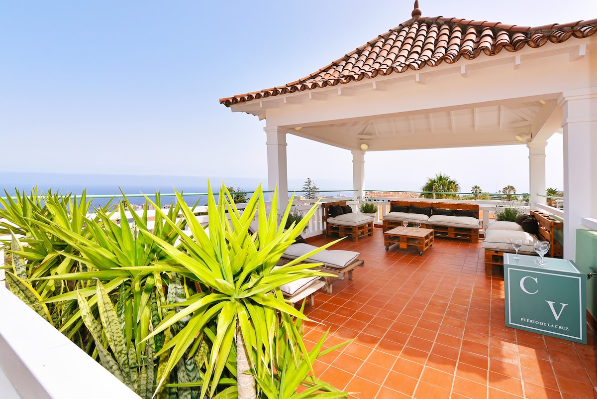 A spacious terrace features a tiled floor and is furnished with lounge sofas. Lush green plants surround the area, providing a serene environment. The open structure is topped with a decorative roof, offering views of the ocean and surrounding landscape.