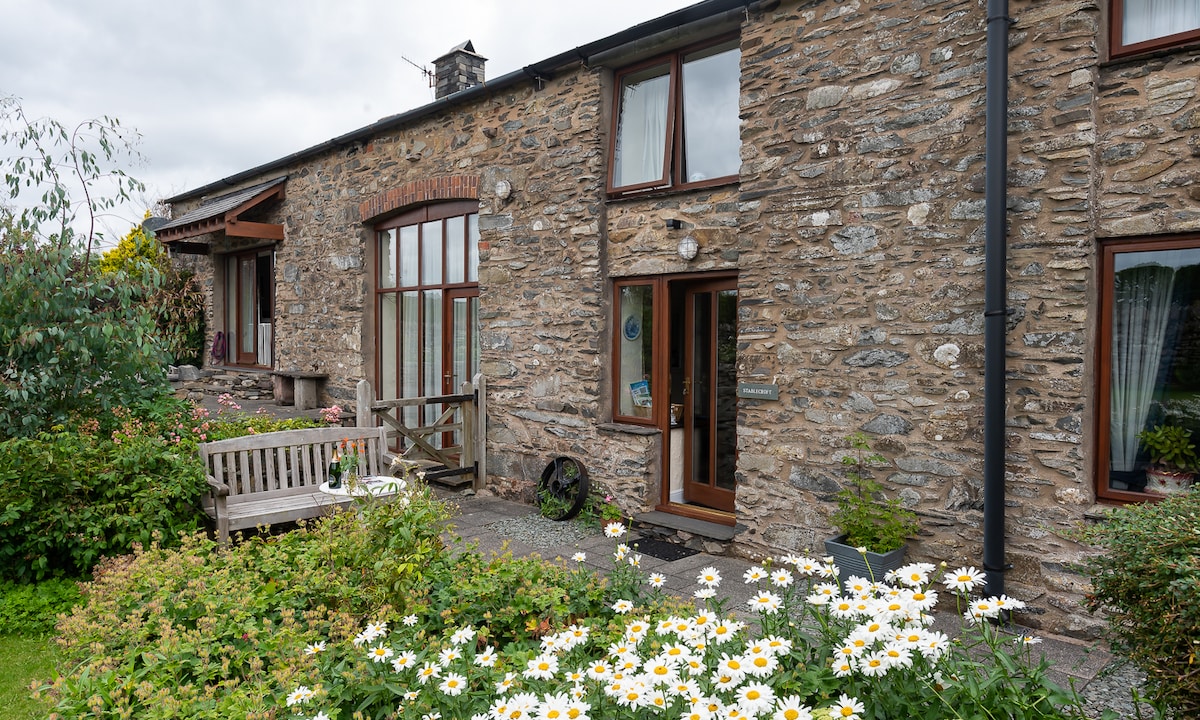The exterior of Stable Croft Cottage is comprised of natural stone walls, featuring large windows that let in ample light. A wooden bench is positioned near the entrance, surrounded by lush greenery and blooming flowers that enhance the tranquil setting.