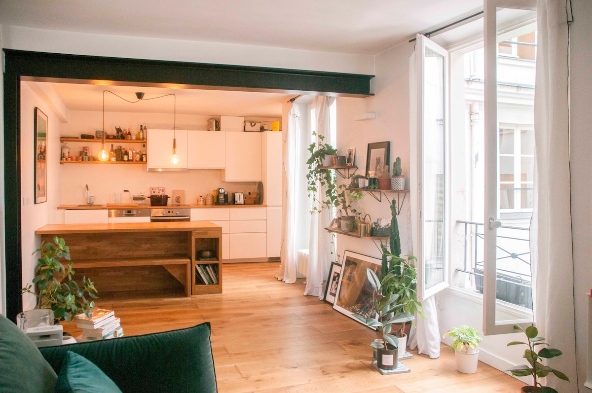 An open kitchen is visible, featuring modern cabinetry and wooden accents. Natural light fills the room through large windows, with plants positioned strategically around. A cozy seating area is observed, complemented by wooden floors and decorative shelves displaying various artistic pieces.