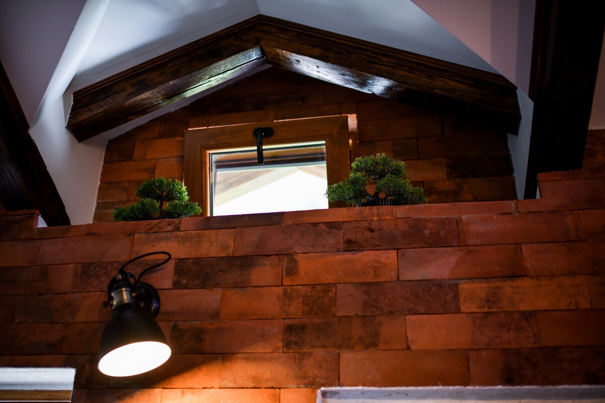 A warmly constructed wall of terracotta bricks features a small window at the top, allowing natural light to filter in. Two potted plants are positioned on the ledge, complemented by a modern black light fixture affixed to the wall, enhancing the interior's character.