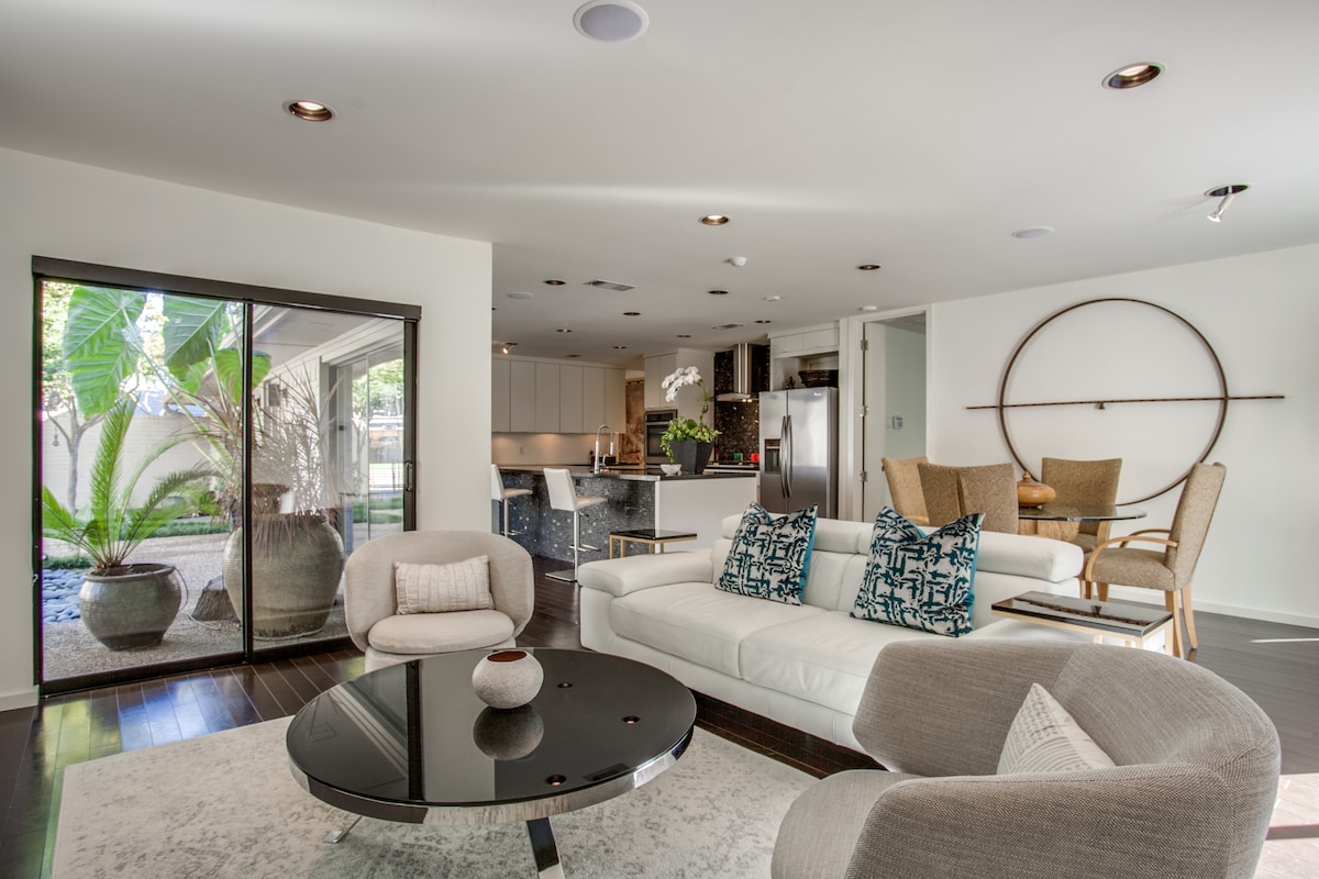 A modern living area is displayed with light-colored sofas and accent chairs. A dark circular coffee table is centered in the space. Sliding glass doors reveal a serene outdoor area with greenery, enhancing the open and airy feel of the home.