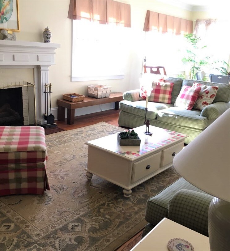 A cozy living area features a fireplace flanked by two seating options. A soft area rug lies beneath a stylish coffee table. Natural light enters through large windows adorned with light-colored draperies, creating a warm ambiance in the space.
