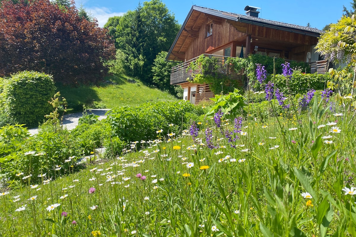 A charming wooden house is surrounded by vibrant greenery and colorful wildflowers. The spacious lawn features blooming daisies and purple flowers, adding a natural touch to the inviting exterior. The deck of the house offers a vantage point to appreciate the beautiful garden and hillside landscape.