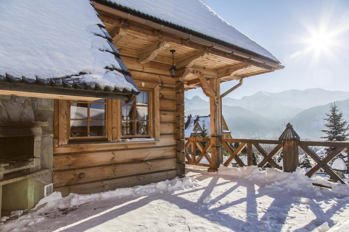 The wooden exterior of the cottage is complemented by a spacious patio featuring a stone BBQ. Snow blankets the ground, and the mountains loom in the background under a bright sun. A wooden railing frames the view, enhancing the rustic charm of the setting.