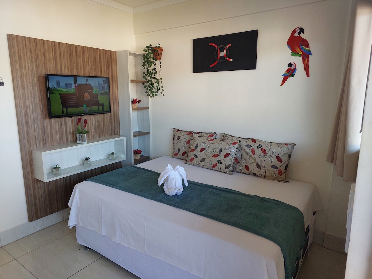 A cozy sleeping area features a spacious bed with white linens and a green throw blanket. Two colorful decorative pillows are positioned against the headboard. A television is mounted on the wall, while a small shelf displays decorative plants. Vibrant artwork and parrot decorations add character to the space.