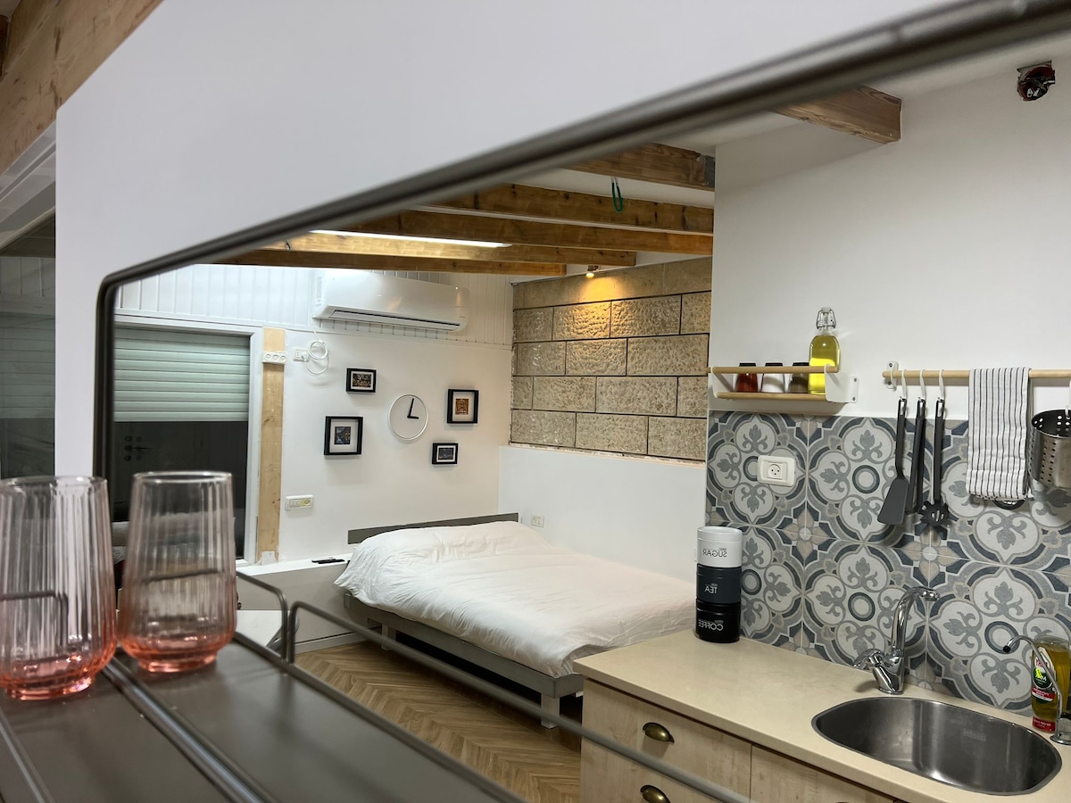 A spacious room is visible, featuring a light-colored bed frame and warm wooden ceiling beams. The reflection in the mirror captures modern kitchen elements, including a sink and cooking utensils, paired with decorative wall art that adds character to the space.