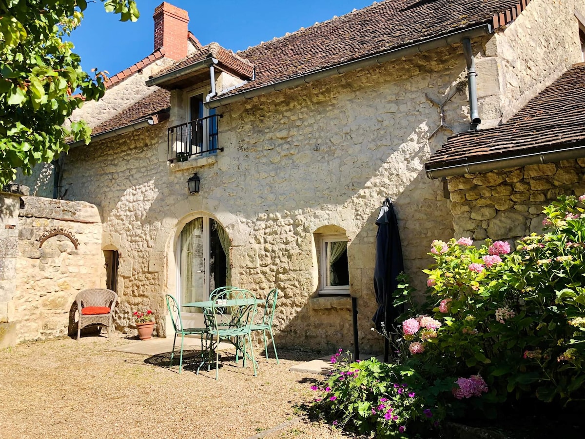 The façade of a charming stone cottage is visible, featuring a mix of light and warm tones. A small table and chairs are set outside among blooming flowers, with a shaded umbrella providing a cozy spot. A balcony can be seen above, adding character to the entrance.