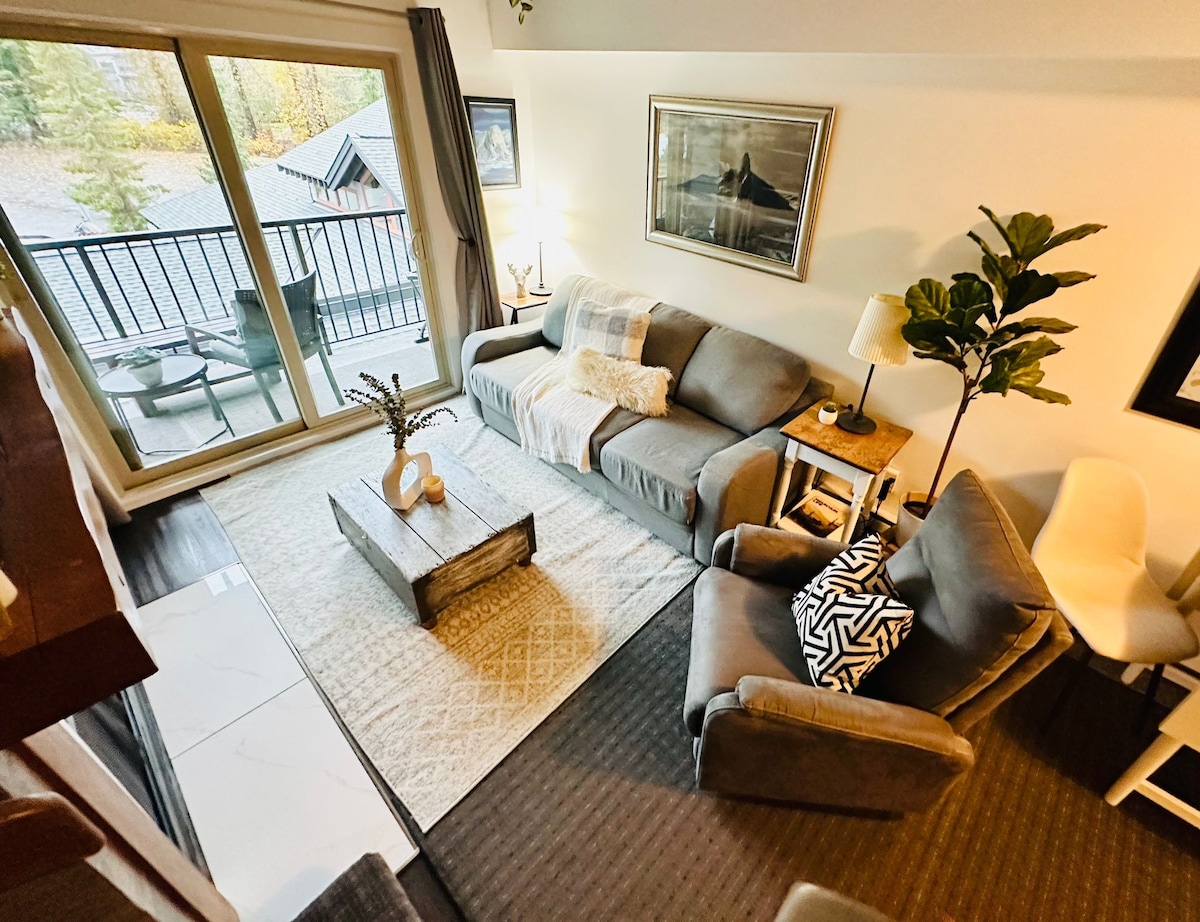 A cozy living area features a gray sofa and armchair arranged around a rustic coffee table, with a warm throw placed casually over the sofa. Large glass doors offer a view of the surrounding trees and access to a private balcony.