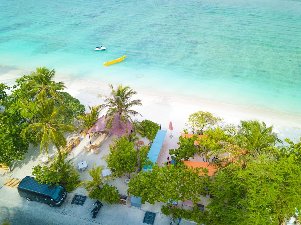 An aerial view showcases a vibrant beachfront area, featuring clear turquoise waters lapping against a sandy shore. Lush palm trees provide shade to the nearby seating areas, while colorful umbrellas and tents add warmth to the inviting outdoor space. Water activities can be seen in the distance.