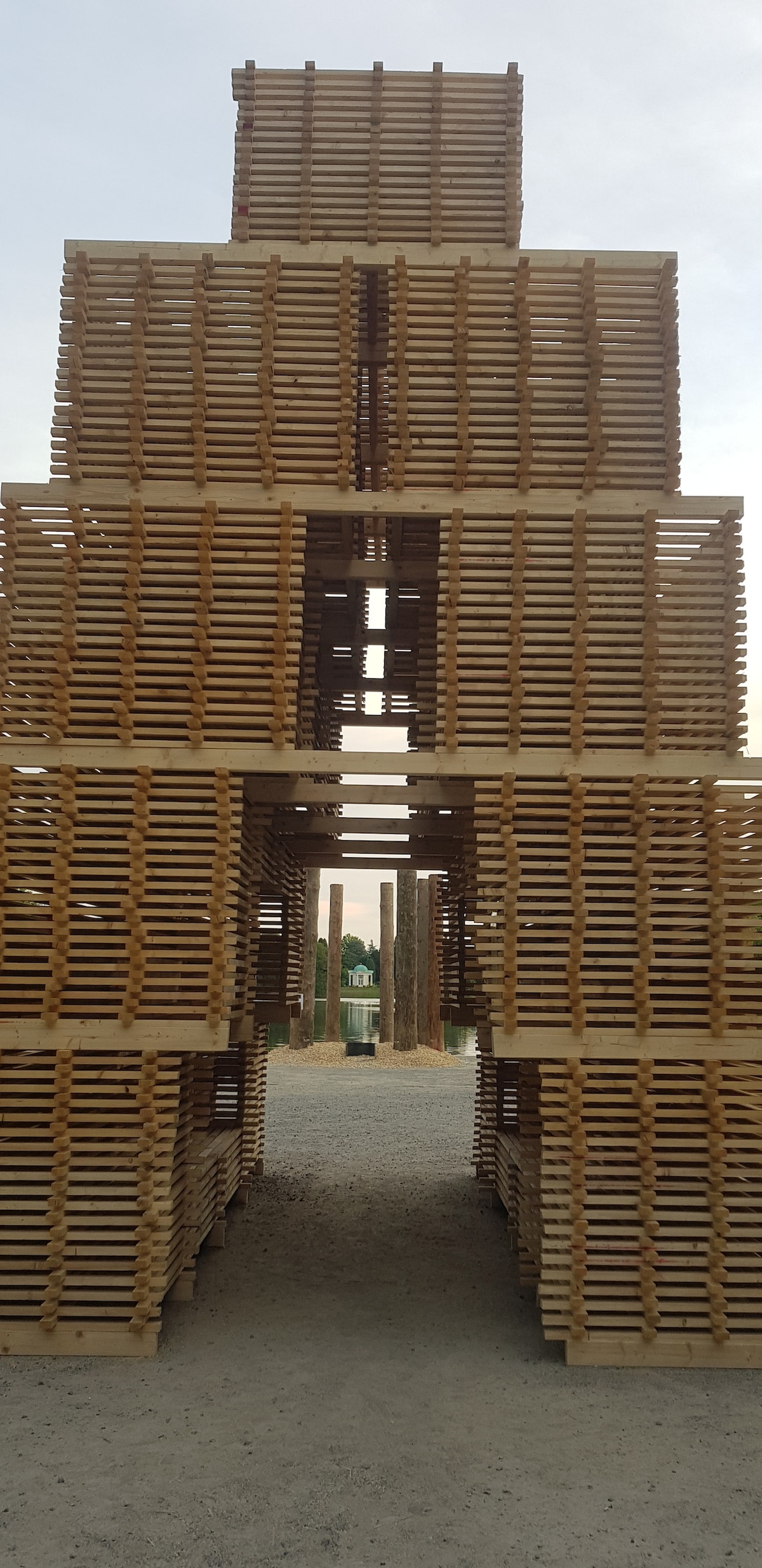 A towering structure made of wooden pallets rises against the sky, creating an interesting architectural formation. Openings between the pallets allow for visibility into the space, with a pathway leading through the stacked arrangement, while the surrounding gravel ground adds a natural element.
