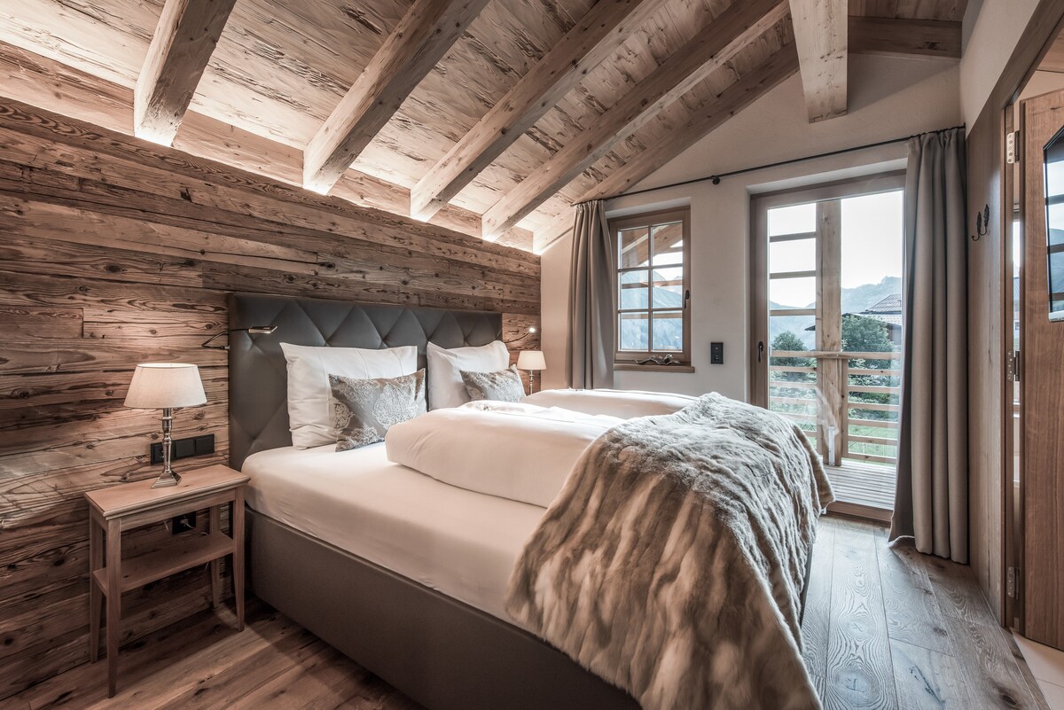 The cozy bedroom features a spacious double bed with a textured blanket. Wooden beams accentuate the ceiling, and natural light streams through a window with views of the surrounding landscape. A bedside table with a lamp adds a warm touch, complemented by wooden wall panels.